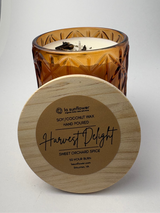 An amber glass jar candle with white wax sits behind a round light wood lid in front of it. The lid has a kraft paper label that reads 'la sunflower' and 'Harvest Delight'.