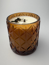 An amber glass candle with a faceted, diamond-cut pattern and white wax, featuring dried botanicals on top, set against a plain light gray background.